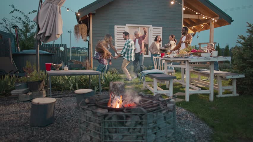 Camera moving closer to positive family. People spending time together. Celebrating something. Everyone dancing. Fire burning at fireplace. Table covered with many dishes. Evening.