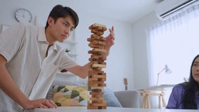 Asian young man and woman playing wood block game in living room at home. Attractive couple players take turn pull a wood block out of the tower and place it on top, enjoy playing board game in house. - Powered by Shutterstock - Get 15% off with code: PIKWIZARD15