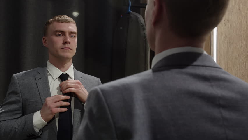 Young businessman in a suit adjusting his tie in front of a mirror, preparing for a business meeting or presentation, concept of self confidence and preparation in the business world