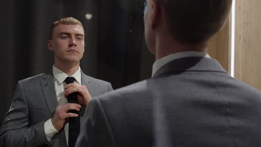 Young businessman in a suit adjusting his tie in front of a mirror, preparing for a business meeting or presentation, concept of self confidence and preparation in the business world