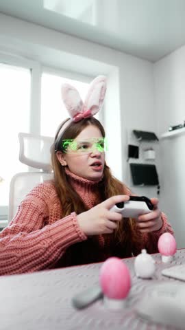 Happy gamer girl in bunny ears and LED glasses enjoys gaming in festive Easter-themed room. She holds controller, celebrating victory while surrounded by Easter eggs and decorations.