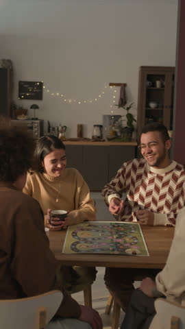 Vertical shot of four friends actively playing board game, gathering around wooden table in cozy, modern apartment