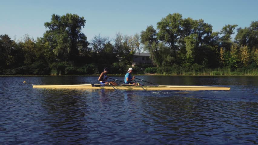 Rowing team training. Side view of 2 young caucasian male rowers, during a rowing practice, athlete sitting in a boat in the river Dnipro, rows through a calm water sunny day, autumn. 4k footage