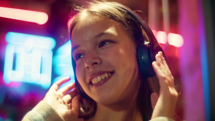 Positive Caucasian Asian teenager daughter girl child dancing junior listening song kid wearing headphones teen enjoy music city evening neon. The inscription noodle in Chinese. Translation: 'noodle'.