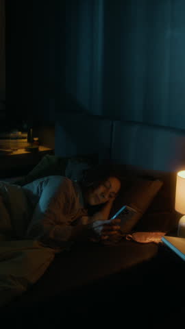 Vertical shot of young woman locking her smartphone and turning off lights before falling asleep in her cozy bed