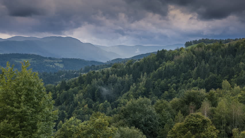 A mountain covered in dense green forests, shrouded in mist under a dramatic sky. Sun rays break through the heavy clouds, casting an ethereal glow over the peaceful valley below