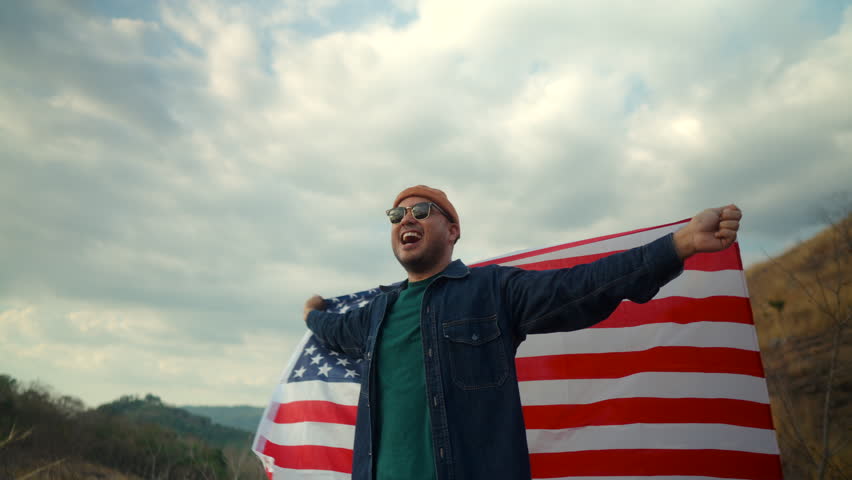 Portrait of cheerful young man wearing sunglasses looking around on the mountain with holding USA flag wegging by wind, proud American holding the United states flag celebration concept.
