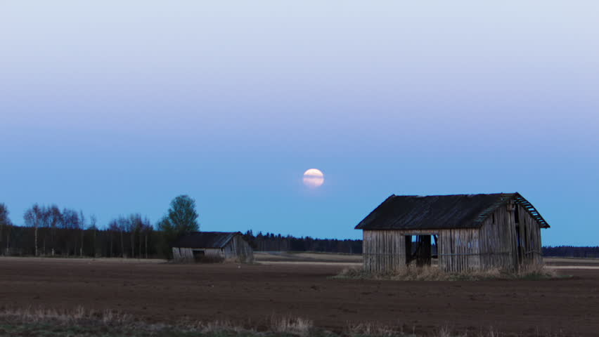 Two old wooden hay barns on a plowed field under a low moon, bird flying across the night sky