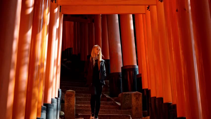 A woman gracefully walks down the stone steps of Fushimi Inari Shrine in Kyoto, Japan, surrounded by countless vermilion torii gates.