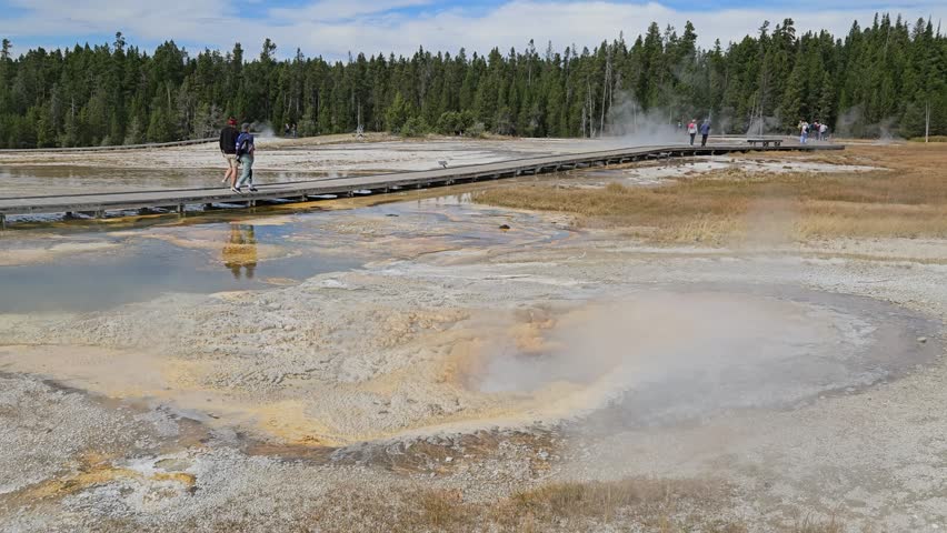 Water and steam is erupting from tiny Geyser. People, tourists are walking on a wooden walkway. Yellowstone National Park, Wyoming, United States