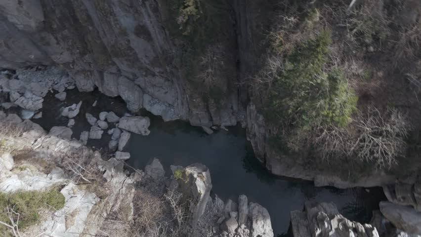 A narrow rocky canyon with steep cliffs and a flowing river below. Large boulders are scattered in the water. Some trees with bare branches grow on the edges, adding depth to the rugged, natural lands