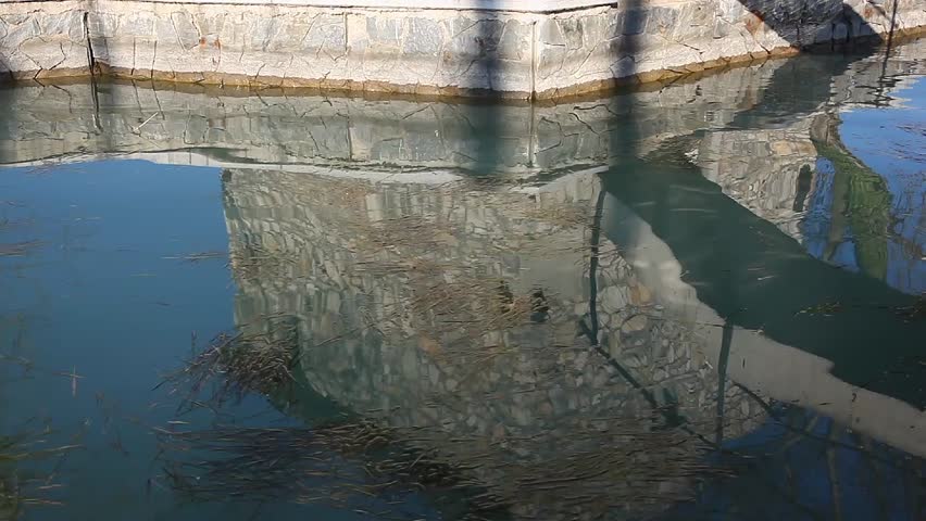 The still water mirrors the stone facade of the house, distorting it in gentle waves. Algae float quietly, while the sun plays with light and shadows on the surface of the small harbor.