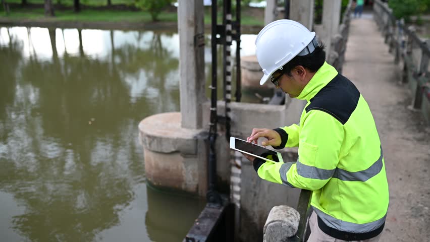 A engineering doing his checking routine. He is wearing hard hat and engineer uniform.Standing by the rail by the dam.Monitor water levels from the heavy rain that has been falling for several days.
