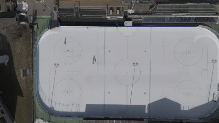 Aerial view of an outdoor ice hockey rink with two skaters casting long shadows. The white ice contrasts with the surrounding area. The rink has markings for hockey games and a spectator stand 