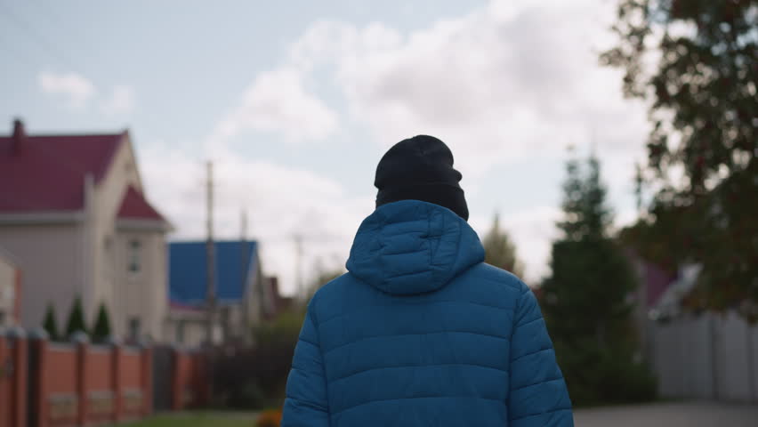 Close up back view of person in blue jacket and black beanie strolling in residential area, with blurred houses and trees in background under cloudy skies