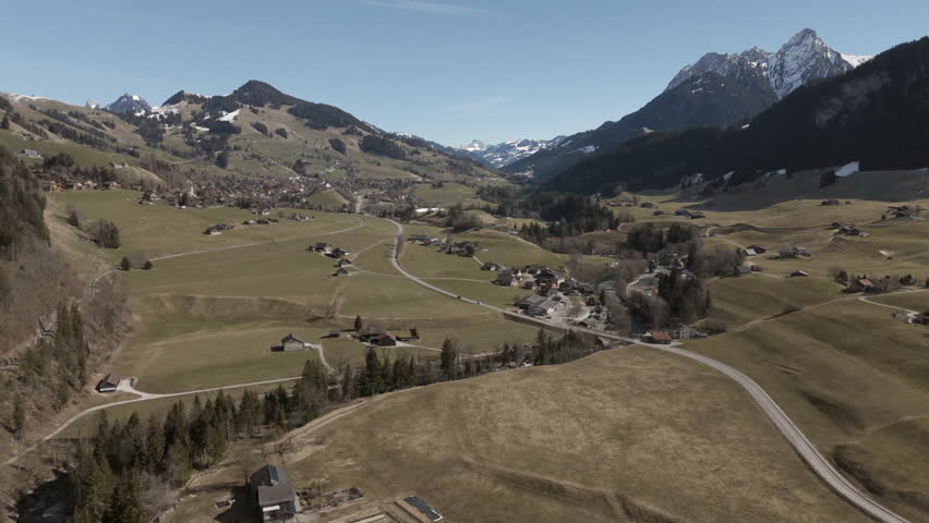 Aerial view of a Swiss alpine village with scattered houses, winding roads, and green fields. Snow-capped mountains rise in the background. A peaceful rural landscape with traditional chalets and natu