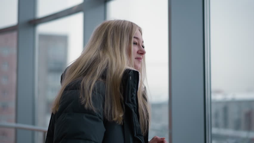 Young woman in black winter jacket looking through glass window with blurred view of cityscape and buildings as she glances behind, feeling relaxed and contemplative in urban environment