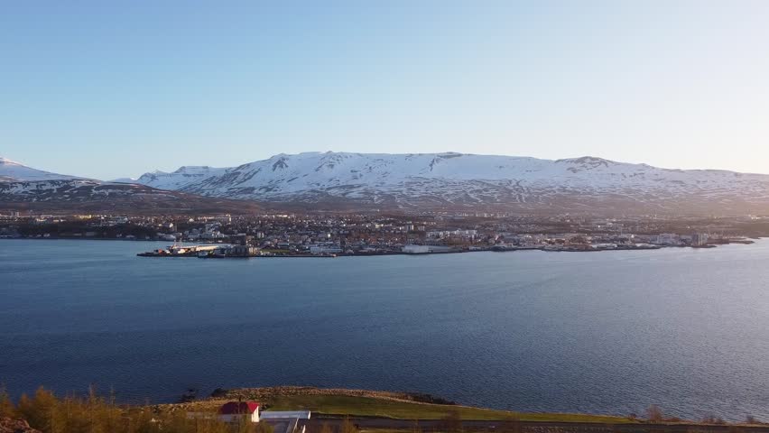 A breathtaking view from Akureyri Hill with calm blue waters and snow-covered mountains. The clear sky highlights the beauty of this Icelandic coastal town.