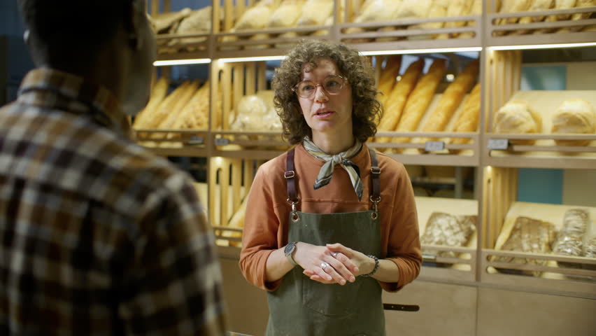 Friendly female bakery worker in apron helping African American customer to choose fresh bread, pointing to assorted loaves on shelves and explaining difference between them