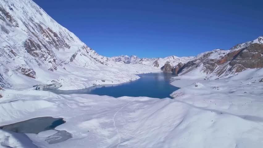 Ascending aerial drone view panorama of Tilicho Lake in Nepal, Annapurna Circuit trekking route. Clear blue glacial lake is surrounded by snowy mountains ranges during sunny day with clear blue sky