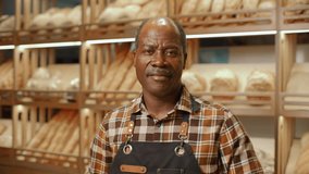 Senior African American baker in apron standing in front of wooden shelves filled with fresh bread in bakery, looking into camera and smiling. Video portrait - Powered by Shutterstock - Get 15% off with code: PIKWIZARD15