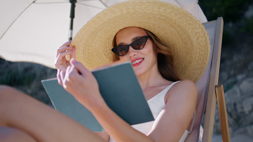 Woman reading book on beach wearing straw hat closeup. Smiling young lady in sunglasses enjoying romantic novel sitting lounger at seashore. Relaxed brunette spending summer sea vacation with hobby.