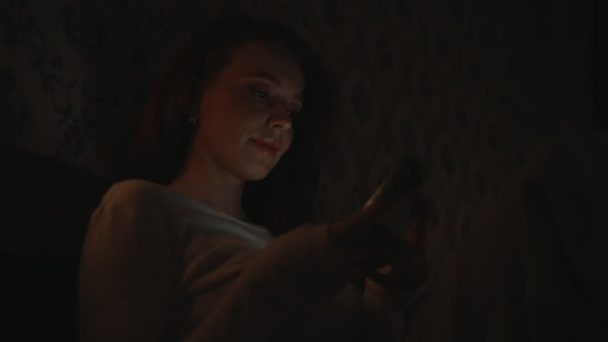Close-up of cheerful young woman relaxing in dark bedroom, scrolling social media on smartphone, screen warm glow illuminating face. Happy redhead female browsing Internet using mobile phone
