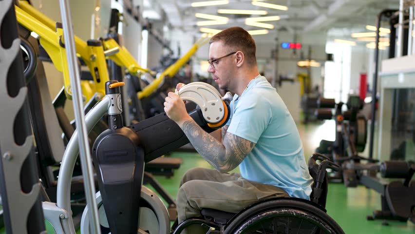 Disabled sportive young man in wheelchair trains upper body muscles, rotating levers in gym. Strong spirit, dedication to sports and health, using arm cycle trainer for rehabilitation
