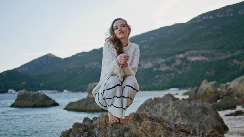 Woman sitting coastal rocks at calm evening. Beautiful relaxed lady squatting on beach stone enjoying morning seascape. Romantic girl relaxing at marine landscape alone. Model posing at sea shore.