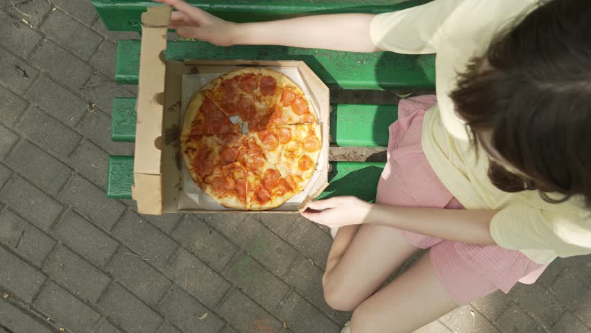 A top-down shot of a teenage girl in a yellow t-shirt and a pink skirt sitting on a bench with a pizza box on it, taking a slice of pepperoni pizza from the box to have a snack on a summer day