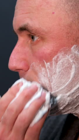 man in robe after shower spa treatments near mirror shaving foam beauty salon self-care at home bathroom bearded handsome man shaving using disposable razor in bathroom looking at mirror.