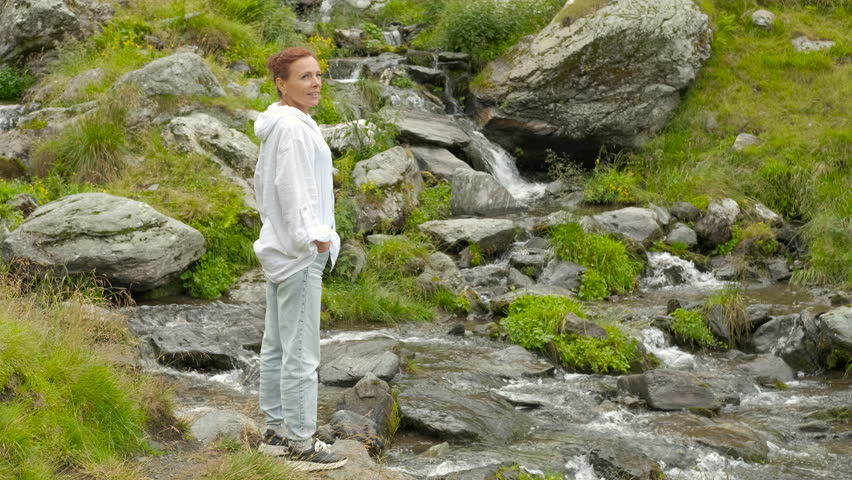 Enjoy the mountain streams. Young hiker is standing near a creek flowing through a rocky landscape. She is looking at the water running between the stones