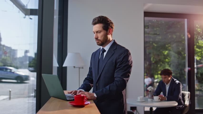Businessman typing laptop in cafeteria enjoying hot coffee. Thoughtful man director creating project working at computer standing cafe counter. Professional entrepreneur surfing online thinking ideas