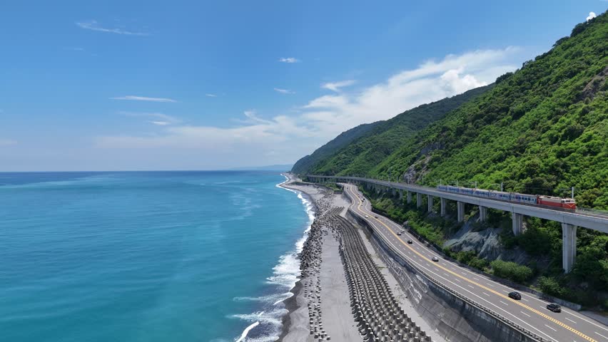 A train  dashing on the elevated tracks of South-Link Railway parallel to a coast highway between green mountains and the azure Pacific Ocean near Duoliang Station in Taitung, Taiwan