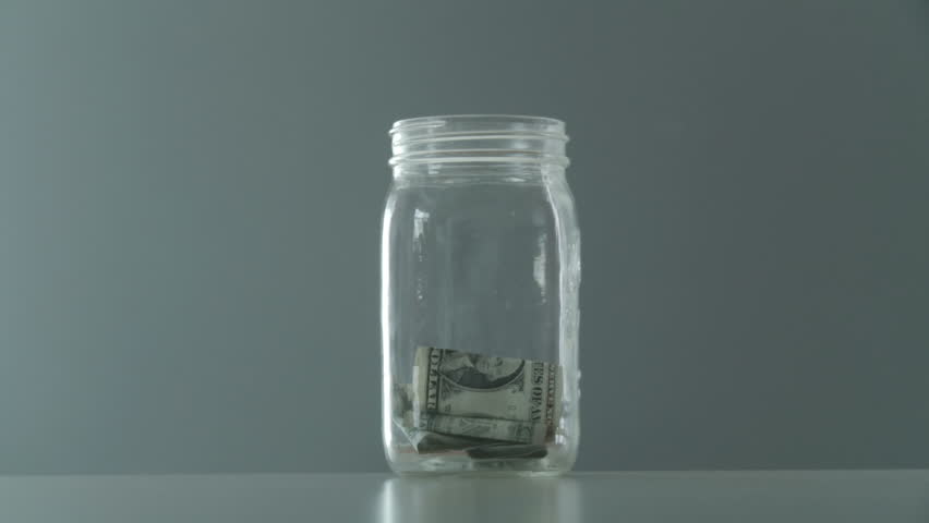 Hand gradually removes dollar and five dollar bills from a glass jar. At one