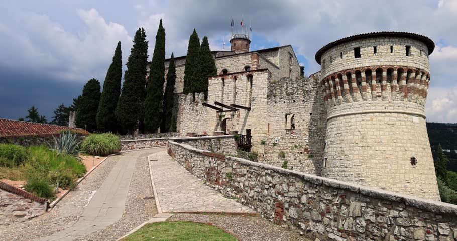 Static video of main entrance to Brescia Castle, showcasing its historic stone walls and fortified tower. Flags of Italy,  European Union,  Lombardy wave atop the main tower under sky