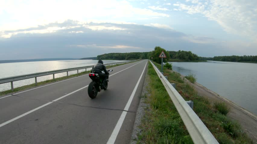 Motorcyclist races his motorcycle on country road at dam. Man in helmet rides on modern sport motorbike at rural route near river or lake. Guy driving bike during trip. Concept of freedom at journey