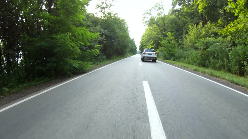 Aerial shot of car riding through forest rural road. White SUV driving at empty country route near wood on summer day. Follow to auto moving during journey or trip. Scenic landscape way. Slow motion