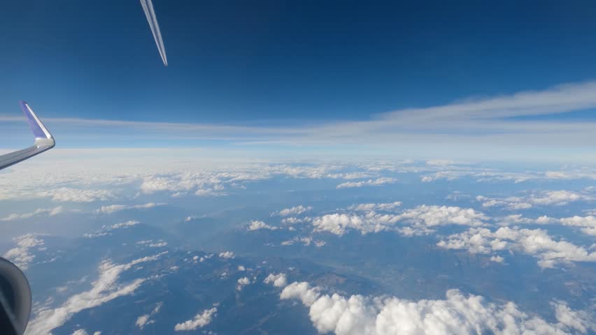 Cumulus clouds over the mountainous terrain and contrail of the other plane, view from an airplane side window during a flight at high altitude
