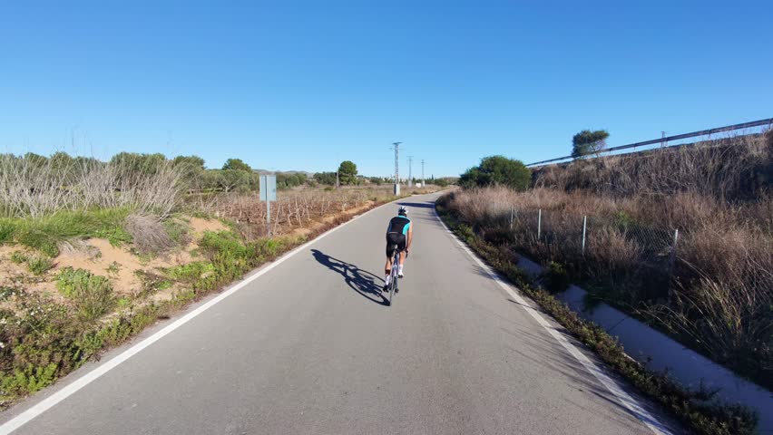 A cyclist rides on a secluded road under a clear blue sky. The sun casts a long shadow. Perfect for themes of fitness, freedom, and outdoor activities.Costa Blanca, Spain