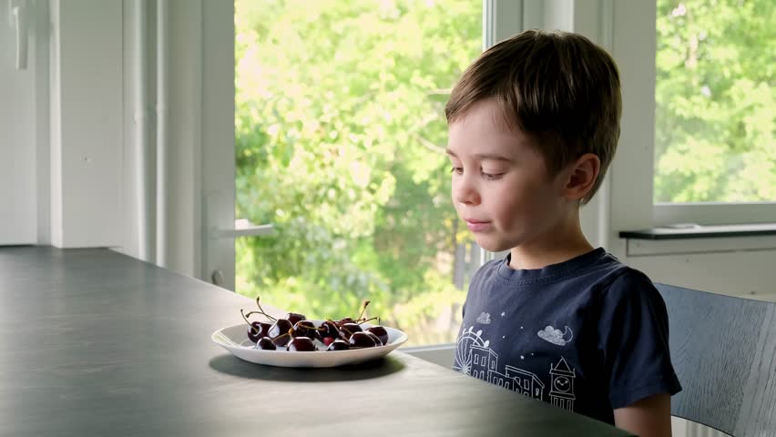 6 year cute little preschool boy eating large ripe cherries. Happy child holding in hand fresh cherry. Healthy organic berries on plate. Summertime season. In the kitchen at the table. Delicious food.