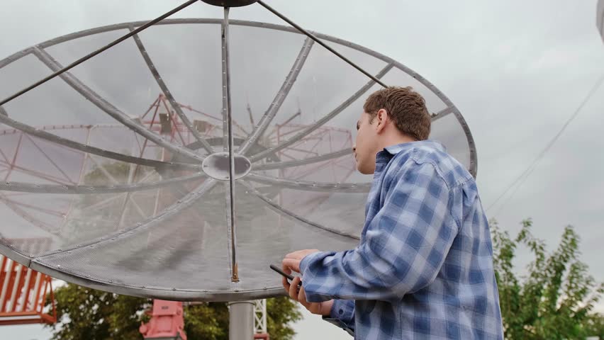 An engineer repairing electronic satellite dishes and radio networks diagnoses issues using a smartphone for troubleshooting.