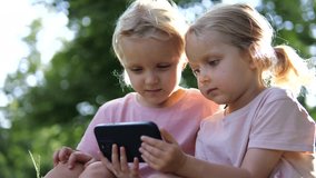 Young blonde sisters closely sharing smartphone screen, sitting side by side on park bench, absorbed in digital content during leisure moment. Concept for children and gadgets - Powered by Shutterstock - Get 15% off with code: PIKWIZARD15