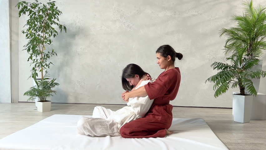 Masseuse making thai yoga massage. Woman therapist doing traditional massage treatment, stretching female patient's body. Client lying on mat, enjoying hand massage in calm atmosphere.