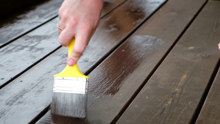 Close-up of a brush painting a wood floor in brown color. Wood plank impregnation. Oil coating of a board. Second layer of oiling. Manual construction work in a gazebo. Natural building materials. 4K.
