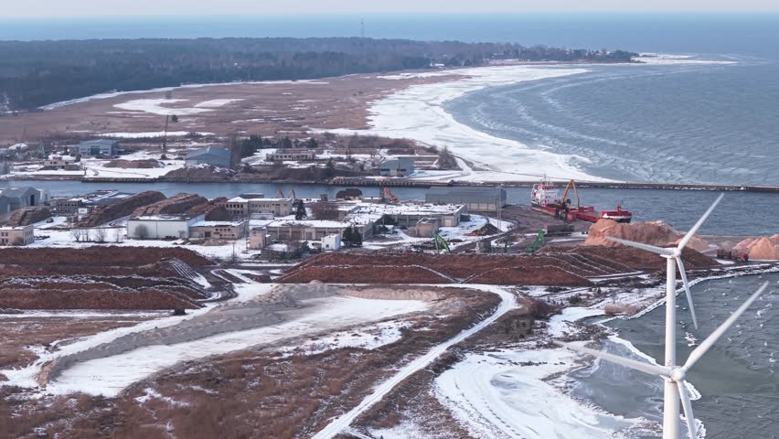 Aerial tracking left telephoto of a timber transshipment port covered in snow, with cranes and shipping vessels in harbor