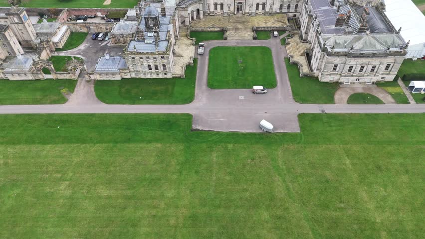 Aerial drone tilt up shot of historic palace with grand architecture, domed roof, and symmetrical wings. Lush gardens, fountains, and pathways extend into distance. Vehicles parked on driveway.