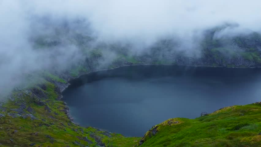 Aerial drone footage of a large blue lake in between Lofoten Norway mountains with white clouds moving between and over the lake low. Green moss is covering the rocky mountains and shoreline at lake.