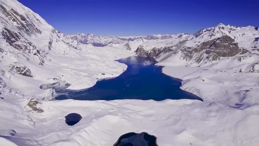 Aerial drone footage flying lower and closer to blue Tilicho lake in Nepal during a sunny day. The lake is surrounded by white covered snowy land and snow covered mountains. Blue sky in the background
