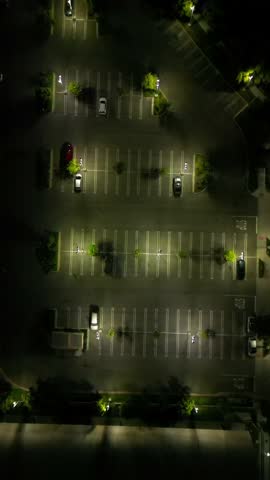 Moody 4K aerial drone footage capturing a quiet parking lot late at night. Features dim streetlights, empty spaces, and occasional parked cars, creating an atmospheric and cinematic urban scene. Perfe